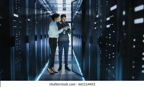 Female IT Technician And Male Server Engineer Talk And Discuss Settings Of A Working Data Center. Man Holds Laptop.