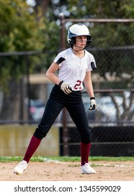 Female Teen Softball Player In Black And White Uniform Taking A Cautious Lead Off From First Base.