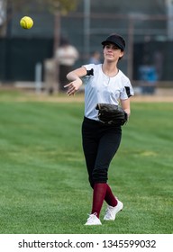 Female Teen Softball Player In Black And White Uniform Throwing Ball On The Outfield Grass.