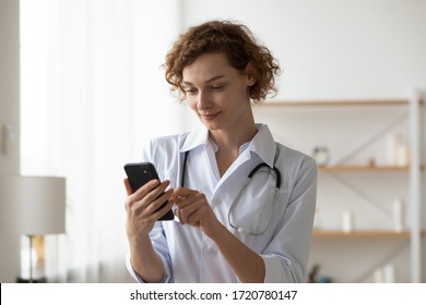 Female Therapist Wearing White Coat And Stethoscope Using Smart Phone App Standing In Clinic. Smiling Lady Physician Holding Mobile Technology Device Texting Message Consulting Remote Patient Online.