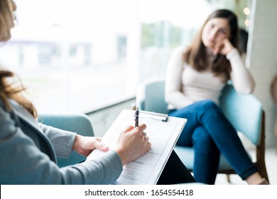 Female Therapist Writing On Notepad While Talking To Patient At Office During Psychology Treatment