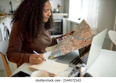 Female Tour Guide Of Black Ethnicity Sitting At Kitchen Table In Her Apartment, Looking At Road Map, Planning New Route For Her Clients, Writing In Her Notepad In Front Of Opened Laptop