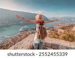 Female tourist with backpack and hat raising arms, celebrating scenic view of Kotor bay from mountain viewpoint at fortress in Montenegro, with cruise ship docked in harbor below