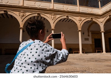 A female tourist captures the beauty of historical architecture in a sunny courtyard using her smartphone, highlighting travel and exploration. - Powered by Shutterstock