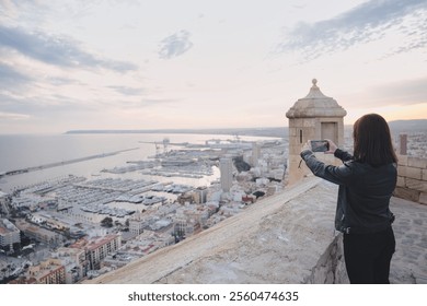 Female tourist capturing the breathtaking panoramic view of alicante cityscape and harbor from the ramparts of santa barbara castle during a beautiful sunset - Powered by Shutterstock