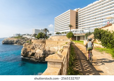 Female tourist enjoying a scenic bike ride along the coast of cales de mallorca, overlooking the clear blue mediterranean sea and a hotel - Powered by Shutterstock