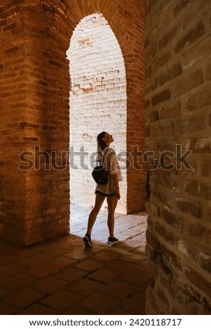 A female tourist gazes up at the towering arches within the brick walls of an ancient fortress bathed in sunlight