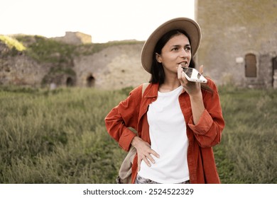 Female tourist in hat dictating voice message into smartphone microphone standing in ancient fortress traveling in Europe. Copy space - Powered by Shutterstock