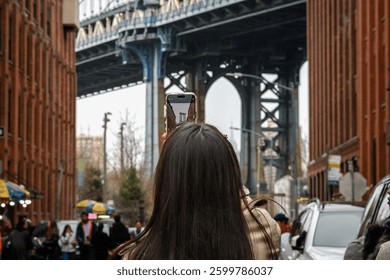 Female tourist taking photo of Manhattan Bridge with her phone in DUMBO, Brooklyn, New York.