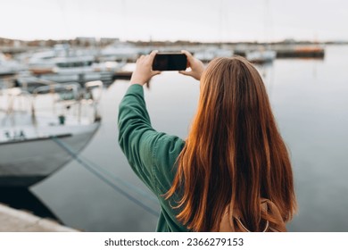 Female tourist taking photo on mobile phone of harbor. Woman walking along the waterfront, seaside. Vacation concept by exploring interesting places to travel