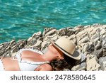 Female tourist wearing sun hat while sunbathing on rocky beach. Red skin and sunburns on chest from not using enough sunscreen.