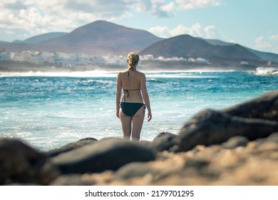 Female tourist at wild rocky beach and coastline of surf spot La Santa Lanzarote, Canary Islands, Spain. La Santa village and volcano mountain in background.