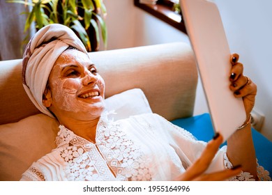 Female With Towel On Hair And Beautician Cream Mask On Face Skin Relaxing Looking At Tablet Computer, Chatting With Friend Online. Laying On Bed At Home, Using Tablet