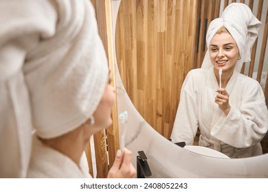 Female with towel turban brushes her teeth in front of mirror