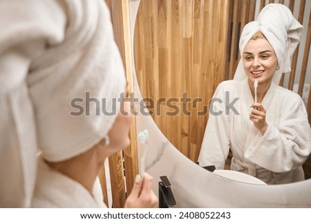 Female with towel turban brushes her teeth in front of mirror