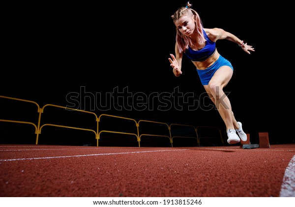 Female track runner bursting off starting\
block against black\
background