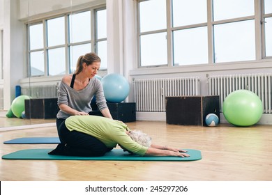 Female trainer helping senior woman doing yoga. Elder woman bending over a exercise mat with personal instructor helping at gym.