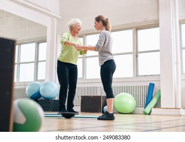 Female trainer helping senior woman in a gym exercising with a bosu balance training platform. Elder woman being assisted by gym instructor while workout session. - Powered by Shutterstock