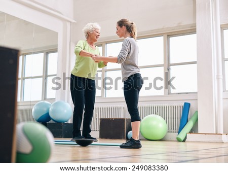 Female trainer helping senior woman in a gym exercising with a bosu balance training platform. Elder woman being assisted by gym instructor while workout session.