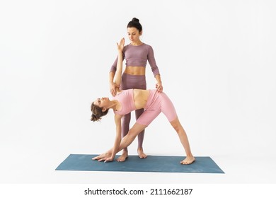 A female trainer helps a student to correctly perform the trikonasana exercise, triangle pose, stand on a mat on a white background