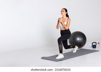 Female Trainer Shows Exercise Lunge With Hands In Front Of Her Chest On Grey Mat And With Gym Equipment In Sport Wear On White Background, Woman Workout With Knee Bent For Stretching