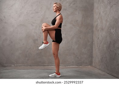 Female trainer in a standing position pulls one leg up to the chest with the knee bent. Young sporty woman doing exercise.