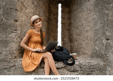 Female travel blogger in a summer dress and straw hat using her laptop while seated in a medieval fortress. Camera and coffee beside her, documenting historical landmarks for her blog. Copy space - Powered by Shutterstock