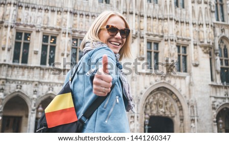 A female traveler with a backpack and a flag of Belgium stands on Grand Place Square in Brussels and shows her thumbs up, Belgium.