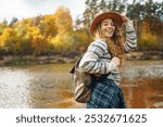 Female traveler with a backpack and a hat enjoying autumn hiking along the river. Active lifestyle. Hiking