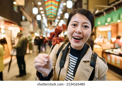 Female Traveler Face To Camera With A Skewer Food Is Excited About Trying An Exotic Snack In Japan. Asian Youtuber Looking At Camera Is Showing Her Fans An Odd Japanese Delicacy Called Taco Tamago.