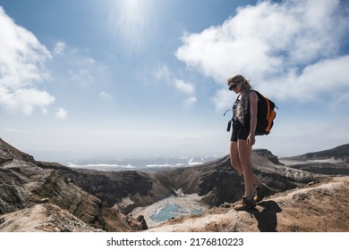 Female Traveler Wearing Sun Glasses And Backpack Walking On Crater Of Volcano Above Blue Lake. Gorely Volcano, Kamchatka