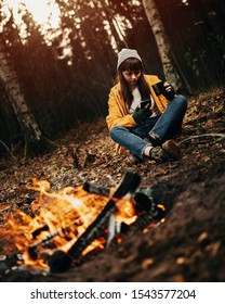 Female Traveler In Yellow Rain Coat Sitting Next To Camp Fire Scrolling Phone And With A Hot Drink In Hand / Dark Autumn Mood In The Woods.