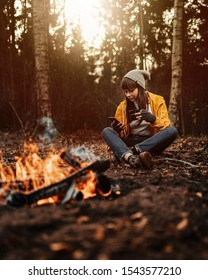 Female Traveler In Yellow Rain Coat Sitting Next To Camp Fire Scrolling Phone And With A Hot Drink In Hand / Dark Autumn Mood In The Woods.