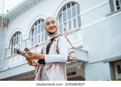 female traveller with hijab smiling while holding the phone and maps with the bulding at the background - Powered by Shutterstock