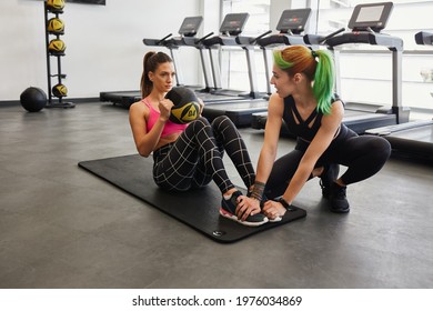 Female Trendy Personal Trainer Holding Young Fit Attractive Female Clients Feet And Assisting During Weighted Ball Ab Crunch Sit Up. Siting On Matt, Gym Equipment In Background