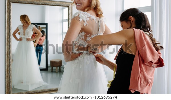 Female\
trying on wedding dress in a shop with women assistant. Female\
assisting bride getting dressed in wedding\
gown.