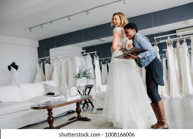 Female Trying On Wedding Gown With Women Assistant In Bridal Wear Shop. Smiling Woman Wearing Her Bridal Dress With Wedding Dress Designer In Bridal Fashion Boutique.