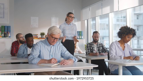 Female Tutor Teaching Class Of Multiethnic Students. Adult Diverse People Sitting At Desk In Classroom Listening To Woman Professor And Writing Down Notes