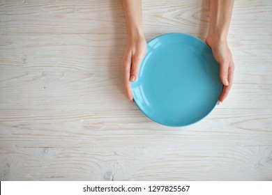 Female two hands hold empty dish on white wooden background. Wooman hands and blue plate top view. Flat lay diet concept.