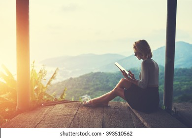 Female is typing something in diary on touch pad, while is sitting against amazing Asian landscape in beautiful summer evening. Woman is watching video on digital tablet during her trip in Thailand