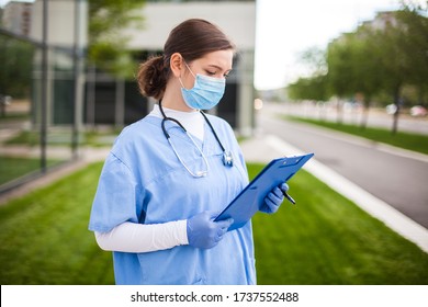 Female UK Doctor Holding Blue Clipboard Standing Outside Hospital Clinic Street Entrance,frontline Key Medical Worker Studying Patient Card Form,modern Care Facility Nursing Home Complex Exterior