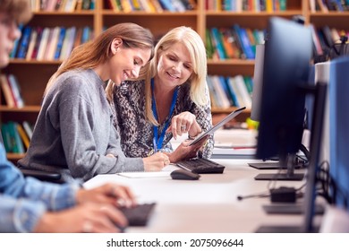 Female University Or College Student Working At Computer In Library Being Helped By Tutor