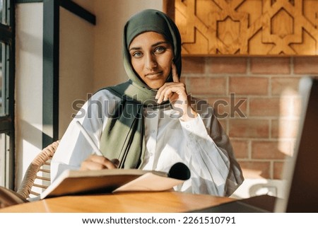 Female university student with a headscarf looking at the camera while writing in a book. Young Muslim woman preparing for an exam while sitting in a campus cafe.