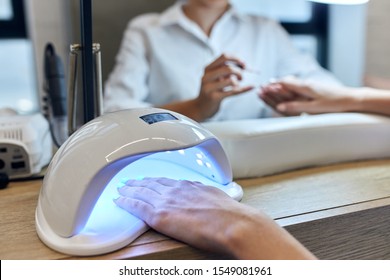female use Ultraviolet lamp or UV in nails salon. Young girl in white clothes drying nails in lamp.