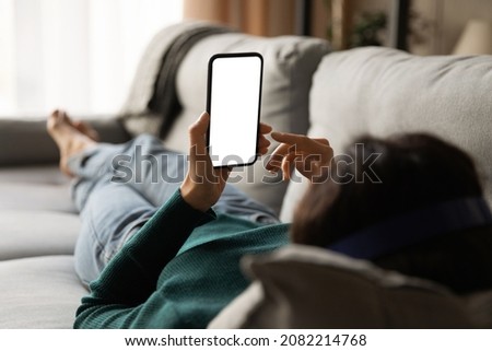 Female using phone. Over shoulder view of young woman lying on sofa hold smartphone with blank empty screen. Template for web app chat interface online advertisement mobile game social network profile