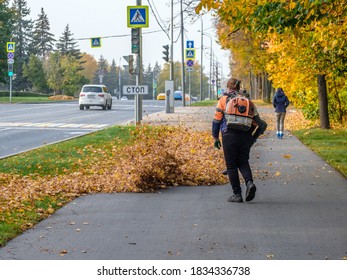 A female utility worker uses a blower to remove fallen leaves in an alley in a park. Yellow leaves are flying in the air. Seasonal work concept.