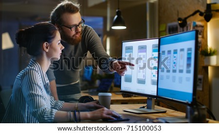 Female UX Architect Has Discussion with Male Design Engineer, They Work on Mobile Application Late at Night. In the Background Wall with Project Sticky Notes and Other Studio Employees.