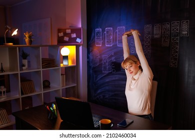 Female UX UI Designer Working Overtime In Home Office Late At Night, Stretching Out While Sitting At Her Desk