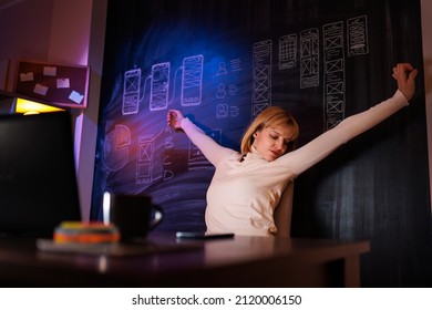 Female UX UI Designer Working Overtime In Home Office Late At Night, Stretching Out While Sitting At Her Desk