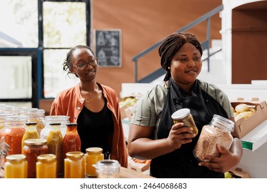 Female vendor and customer walking towards checkout counter with jars of preserved chemical free products. Black woman with an apron carrying bio organic items, assisting client at local store. - Powered by Shutterstock